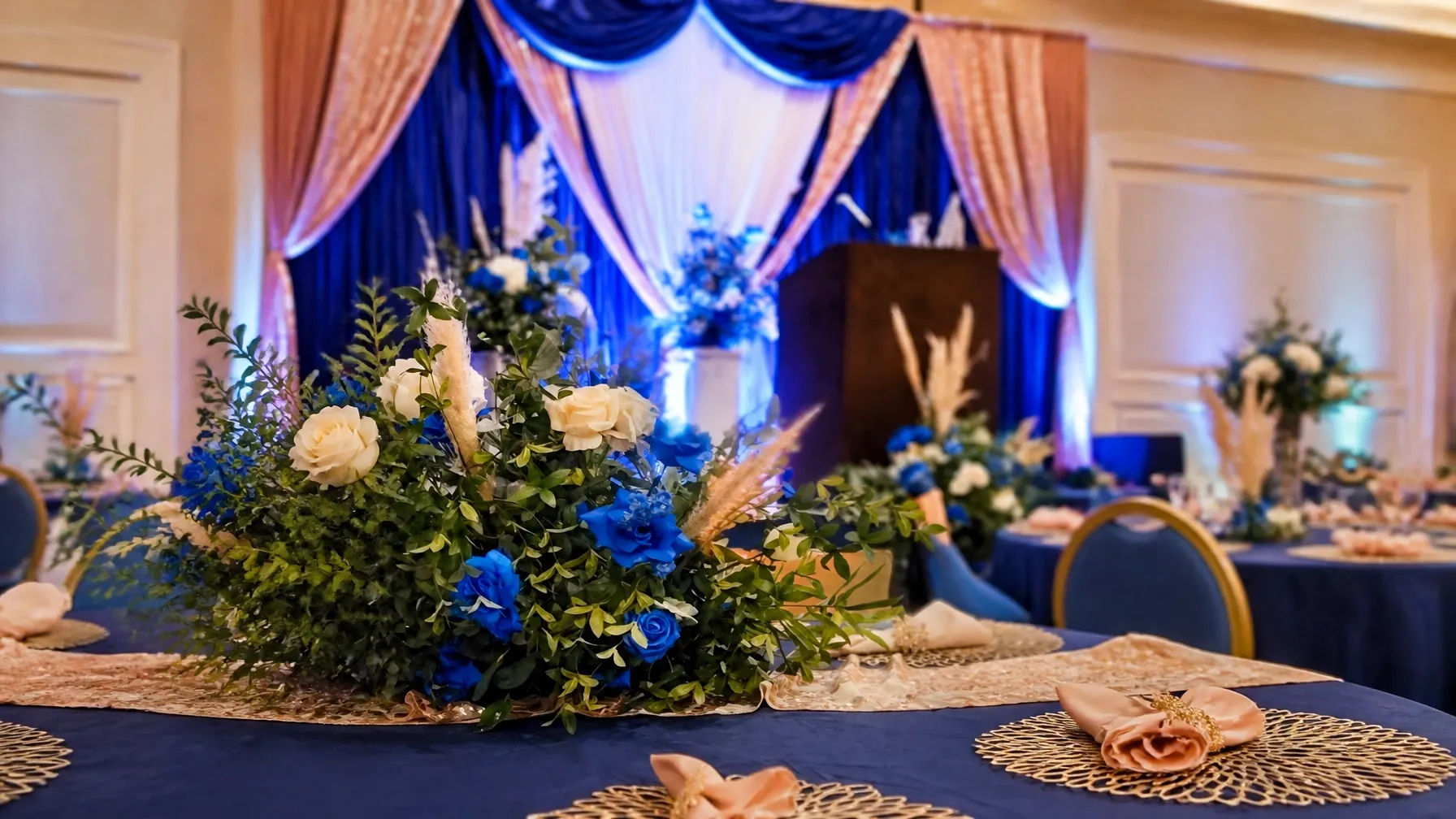 Royal-blue and champagne ballroom setup with draped fabric, floral centerpieces, and layered tables